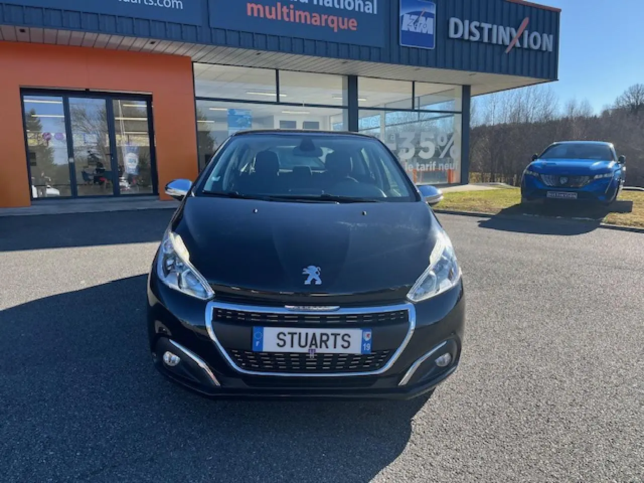 Vue avant d'une Peugeot 208 noire garée devant un bâtiment commercial avec calandre chromée et phares allumés.