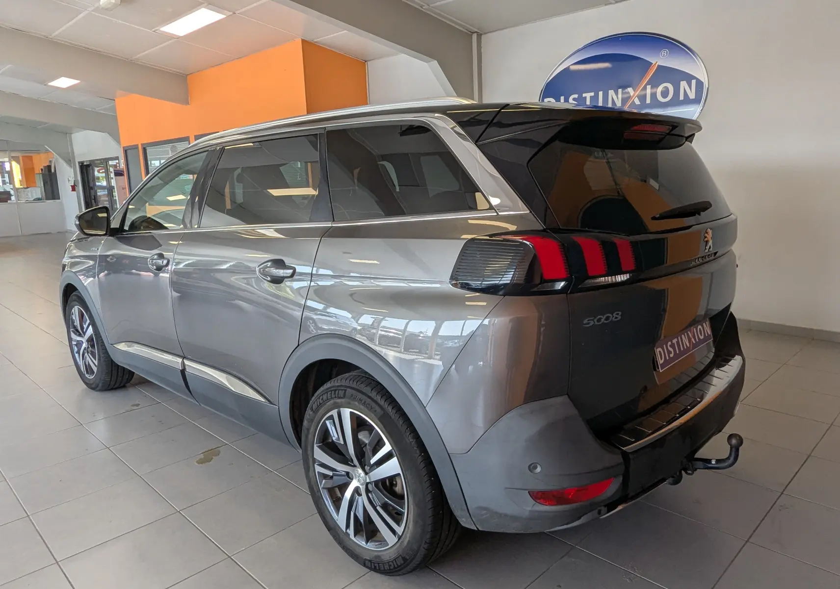 Peugeot 5008 gris vue 3/4 arrière droit en intérieur, avec feux arrière à griffes rouges et attelage visible.