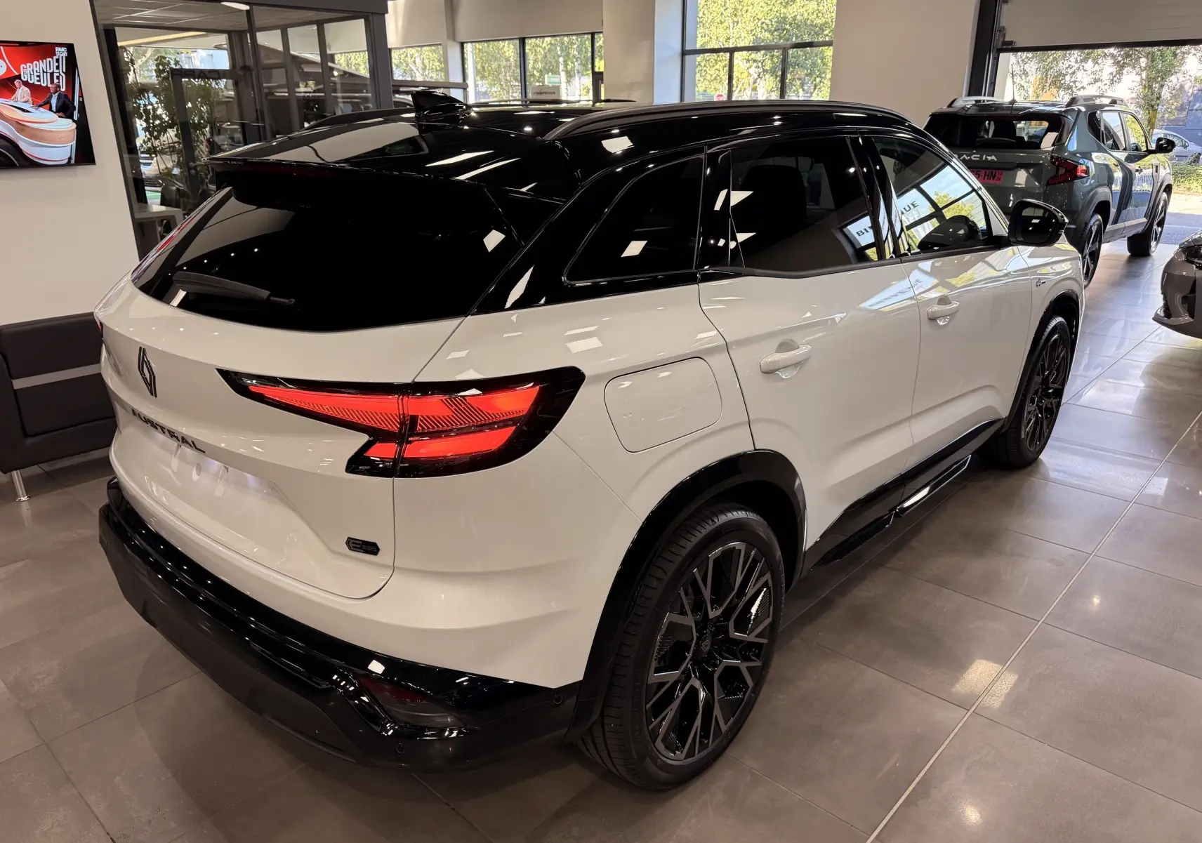 Vue 3/4 arrière droite du Renault Austral blanc nacré avec toit noir, jantes noires et feux arrière LED allumés en showroom.