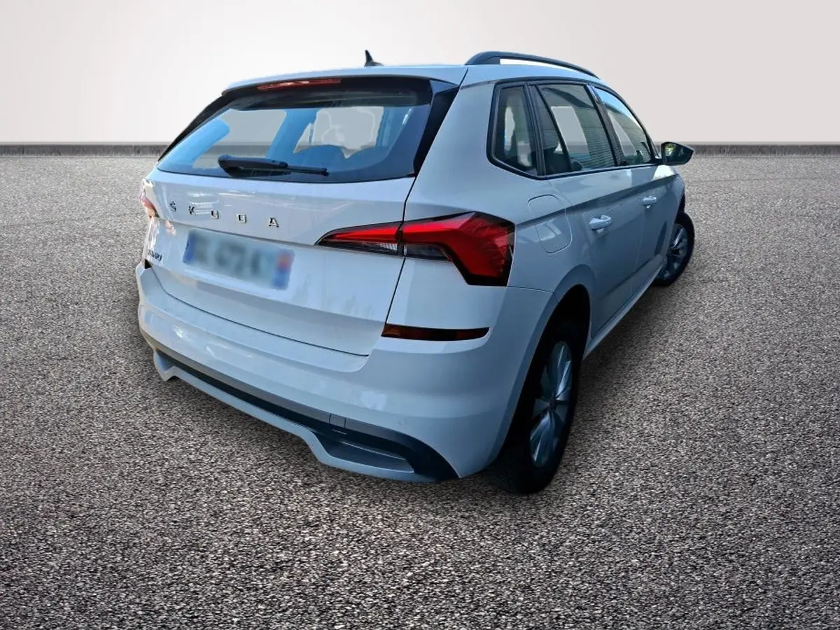 Vue 3/4 arrière droite d'un Skoda Kamiq blanc avec feux LED et barres de toit noires sur sol gravillonné.