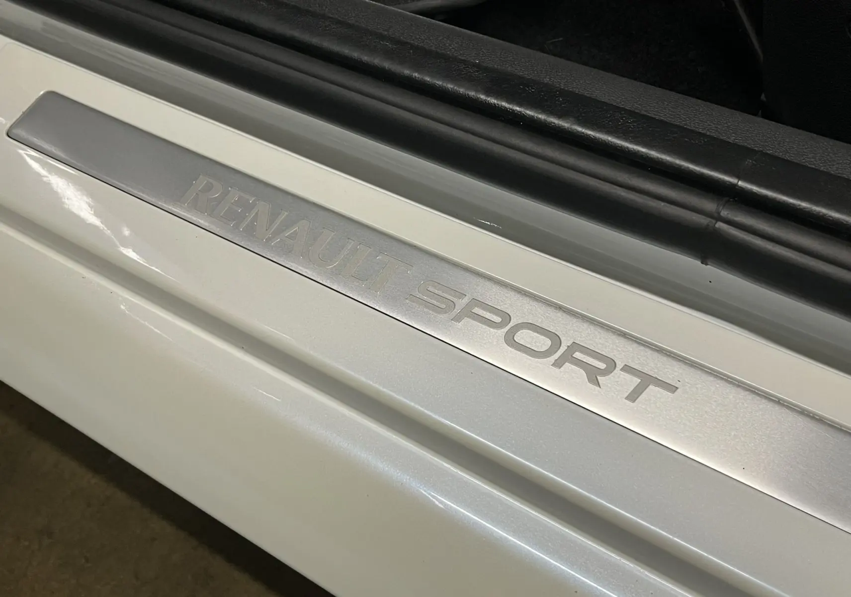 Gros plan sur la plaque de seuil de porte argentée "RENAULT SPORT" sur une Renault Clio III blanc nacré.