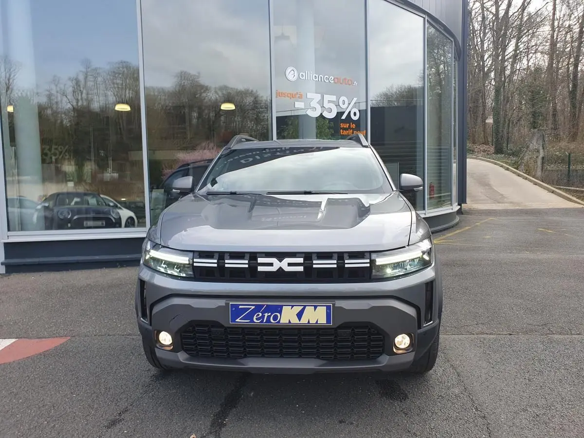 Vue avant d'un Dacia Duster 1.6 Hybrid gris schiste avec calandre noire et feux LED allumés, stationné devant un bâtiment vitré.