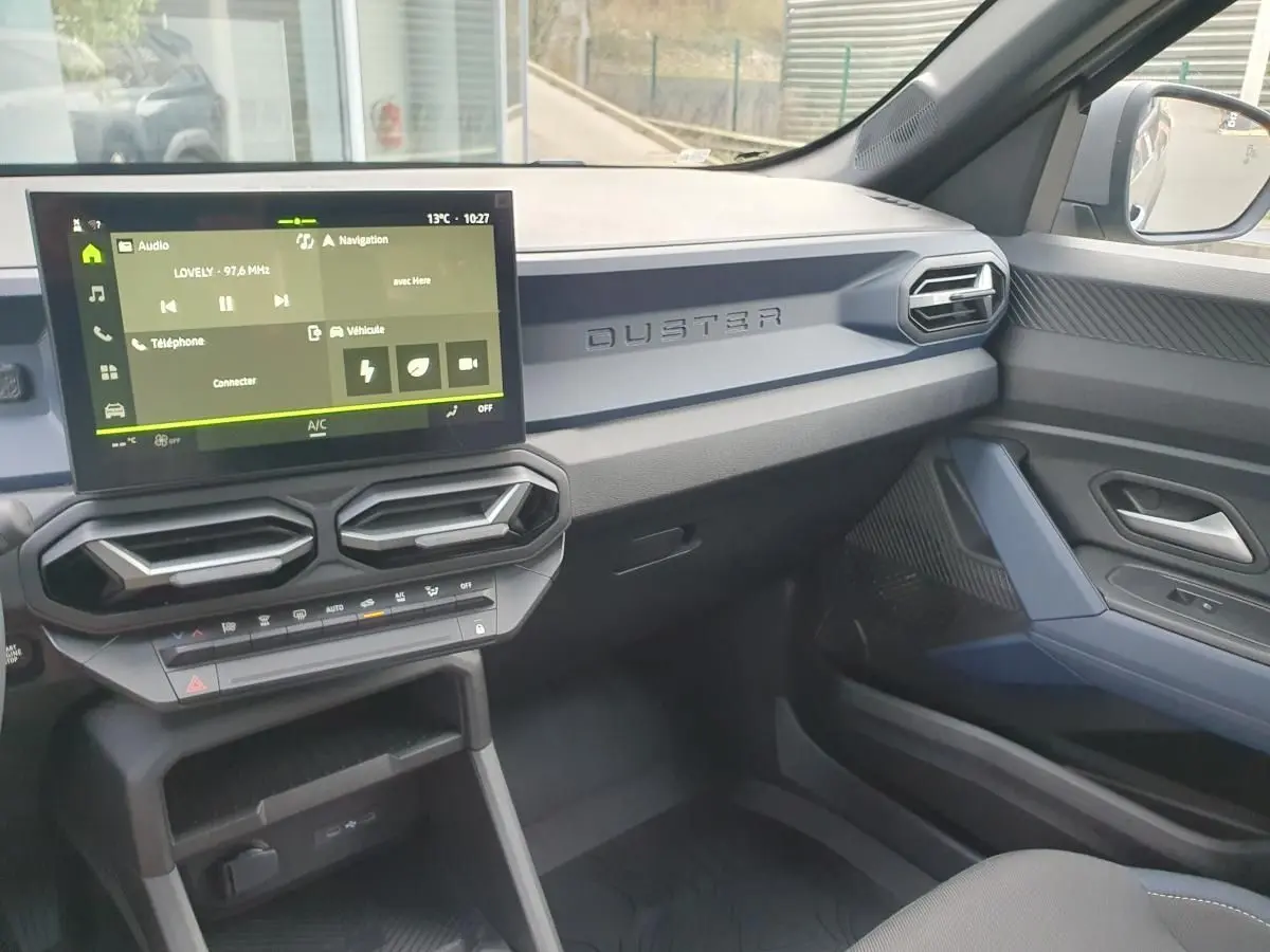 Vue intérieure côté passager du tableau de bord gris du Dacia Duster 2026 avec écran tactile et aérateurs hexagonaux.