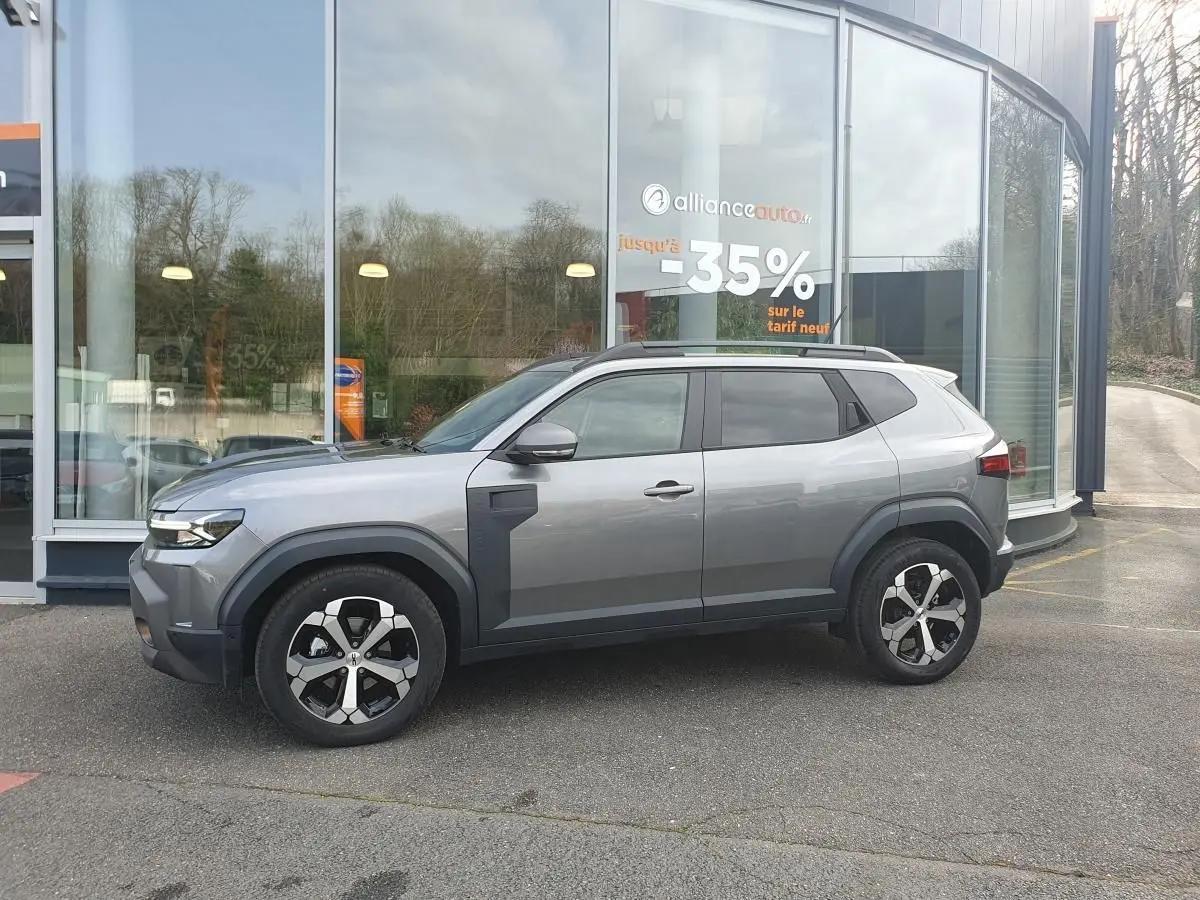 Vue de profil côté gauche du Dacia Duster gris schiste 2026 avec jantes alliage 18 pouces et barres de toit noires.