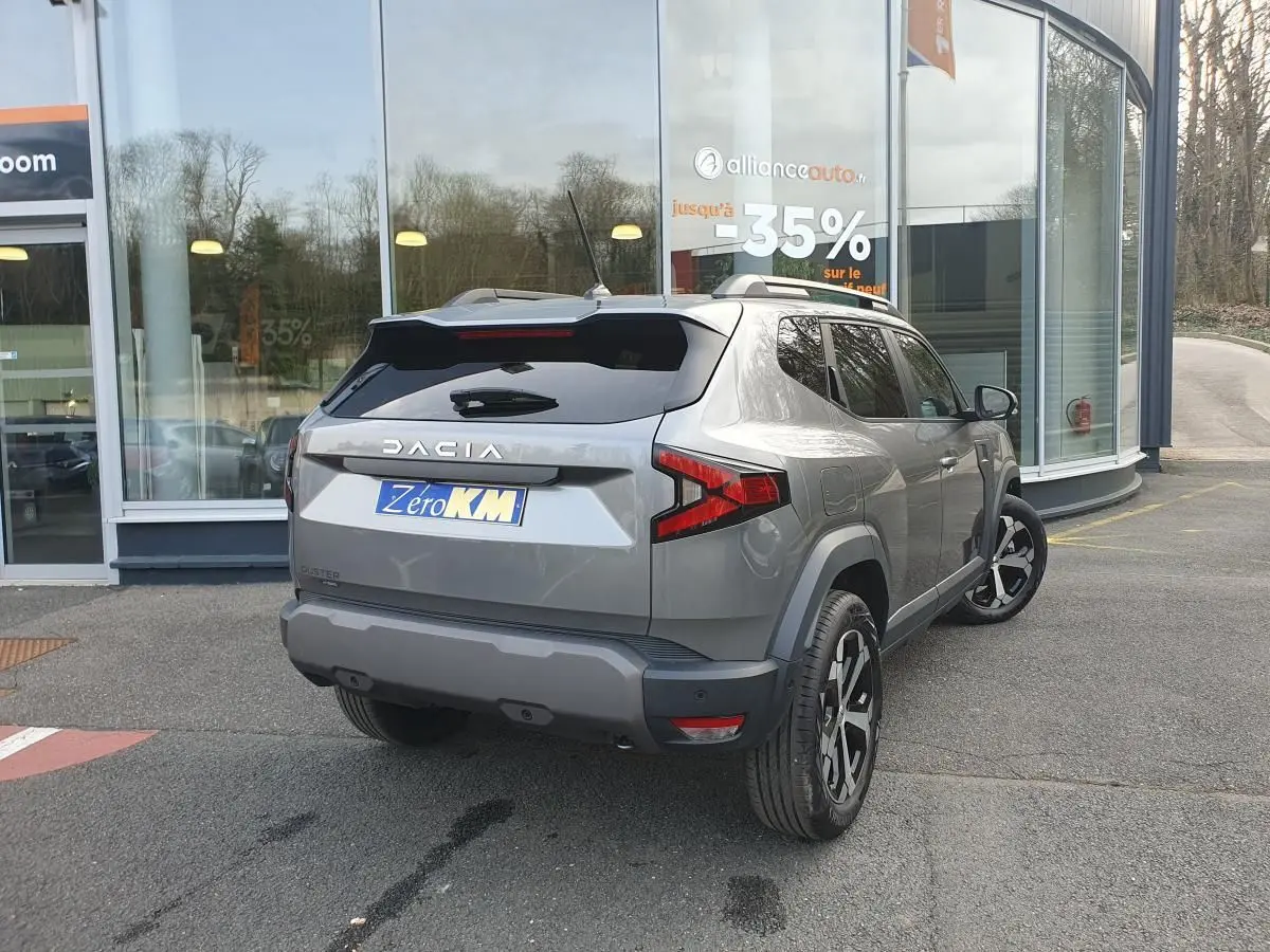 Vue 3/4 arrière droite d’un Dacia Duster gris schiste avec jantes alliage 18” diamantées, stationné devant un showroom.