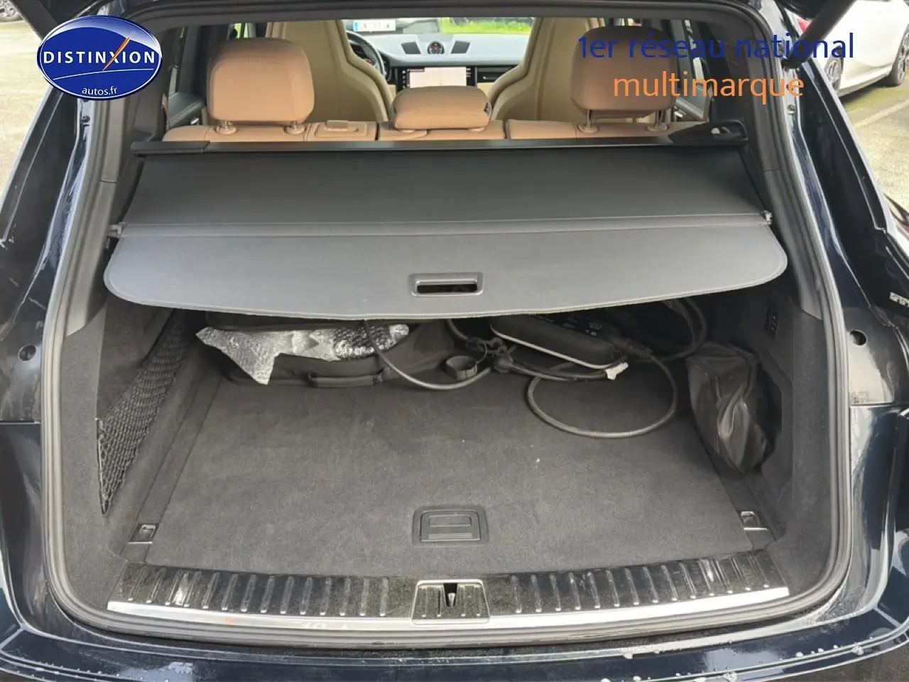 Coffre ouvert d’un Porsche Cayenne bleu, vue arrière, avec cache-bagages et câbles de recharge visibles.
