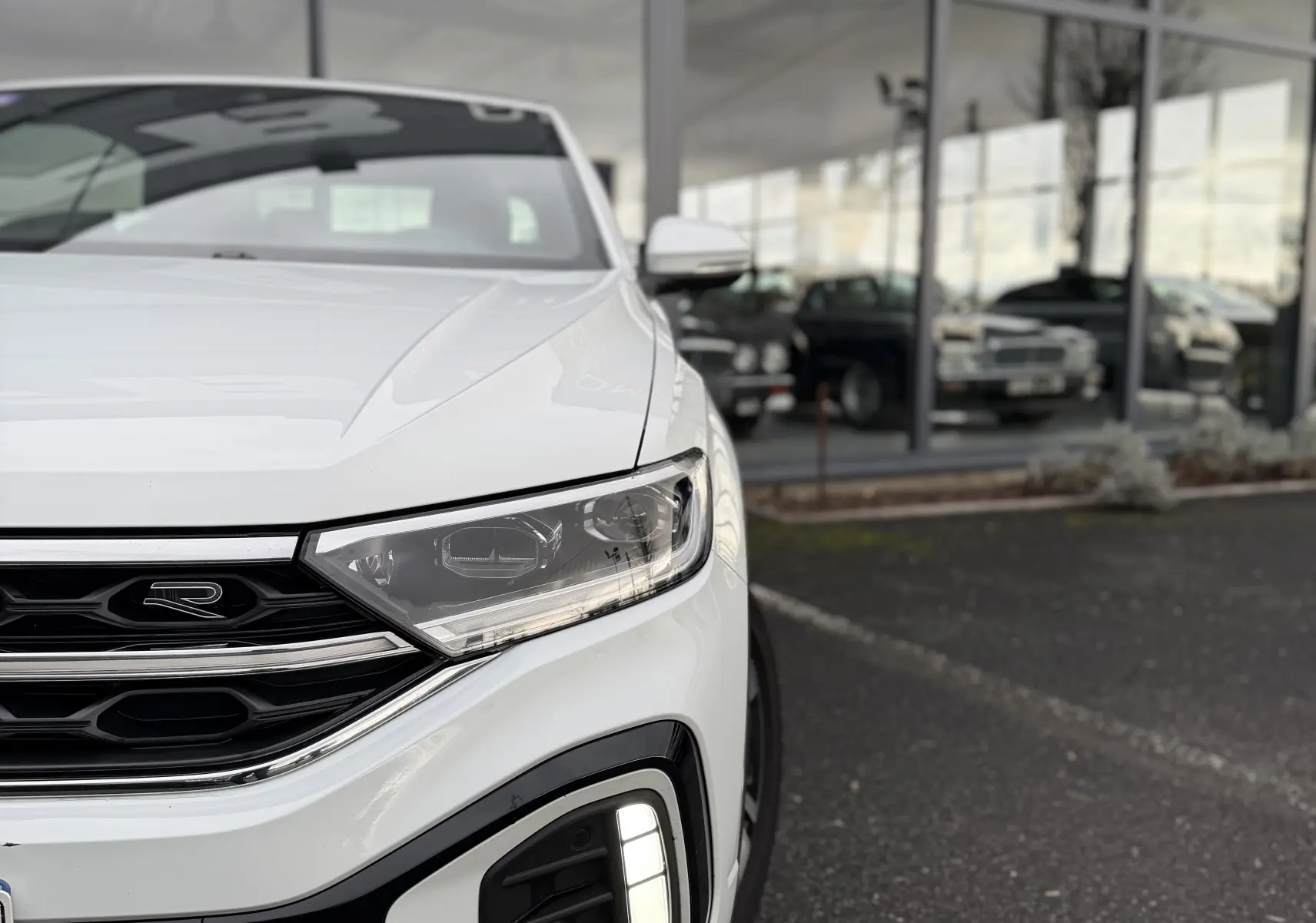 Vue 3/4 avant droit du Volkswagen T-Roc Cabriolet blanc, mettant en valeur le logo R-Line et les feux LED distinctifs.