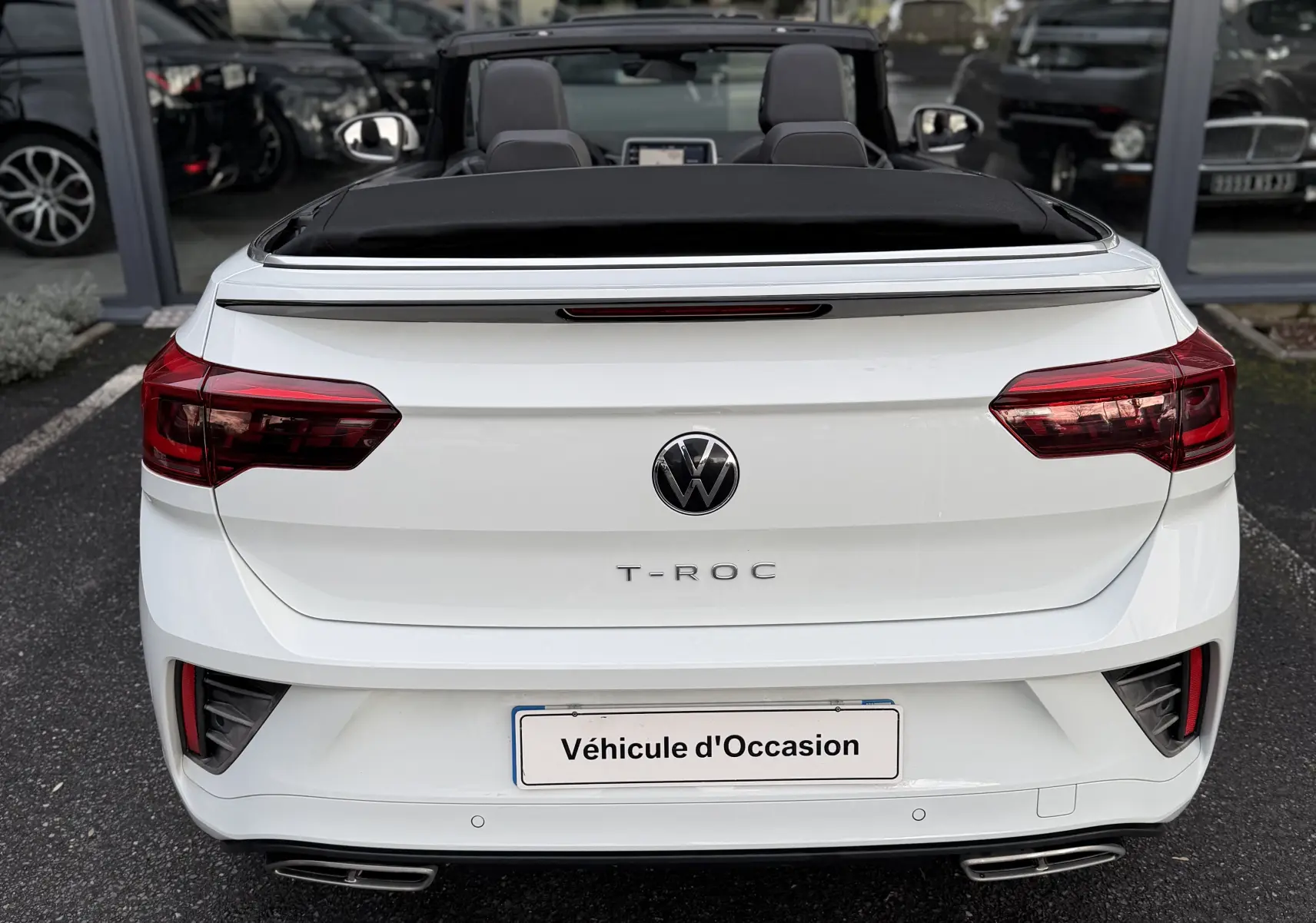 Vue arrière d'un Volkswagen T-Roc Cabriolet blanc avec toit décapoté et logo VW bien visible, stationné devant une vitrine.