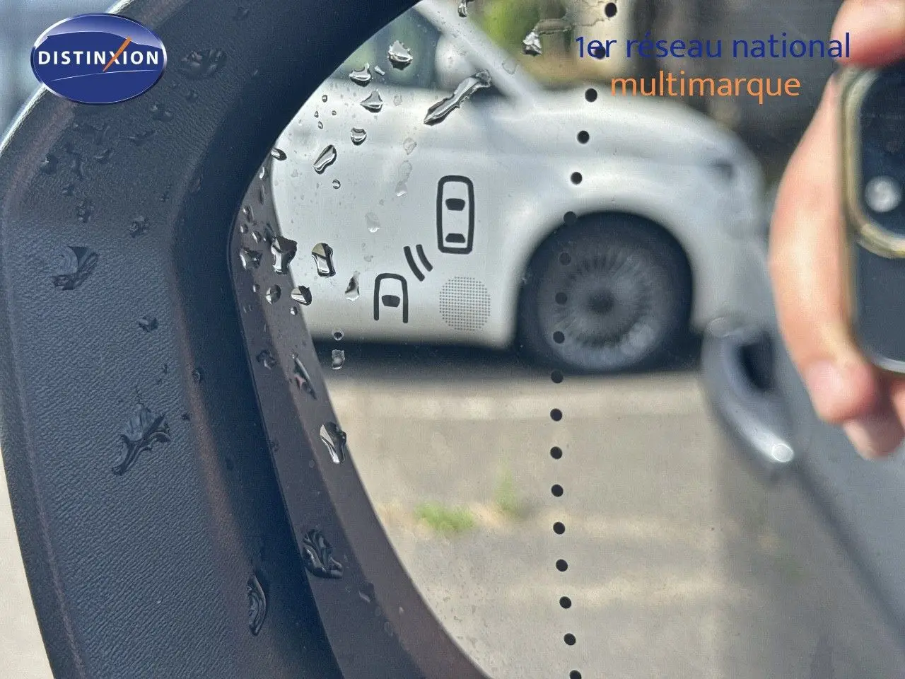 Reflet dans le rétroviseur côté gauche montrant une voiture blanche floue avec gouttes d'eau sur le miroir.