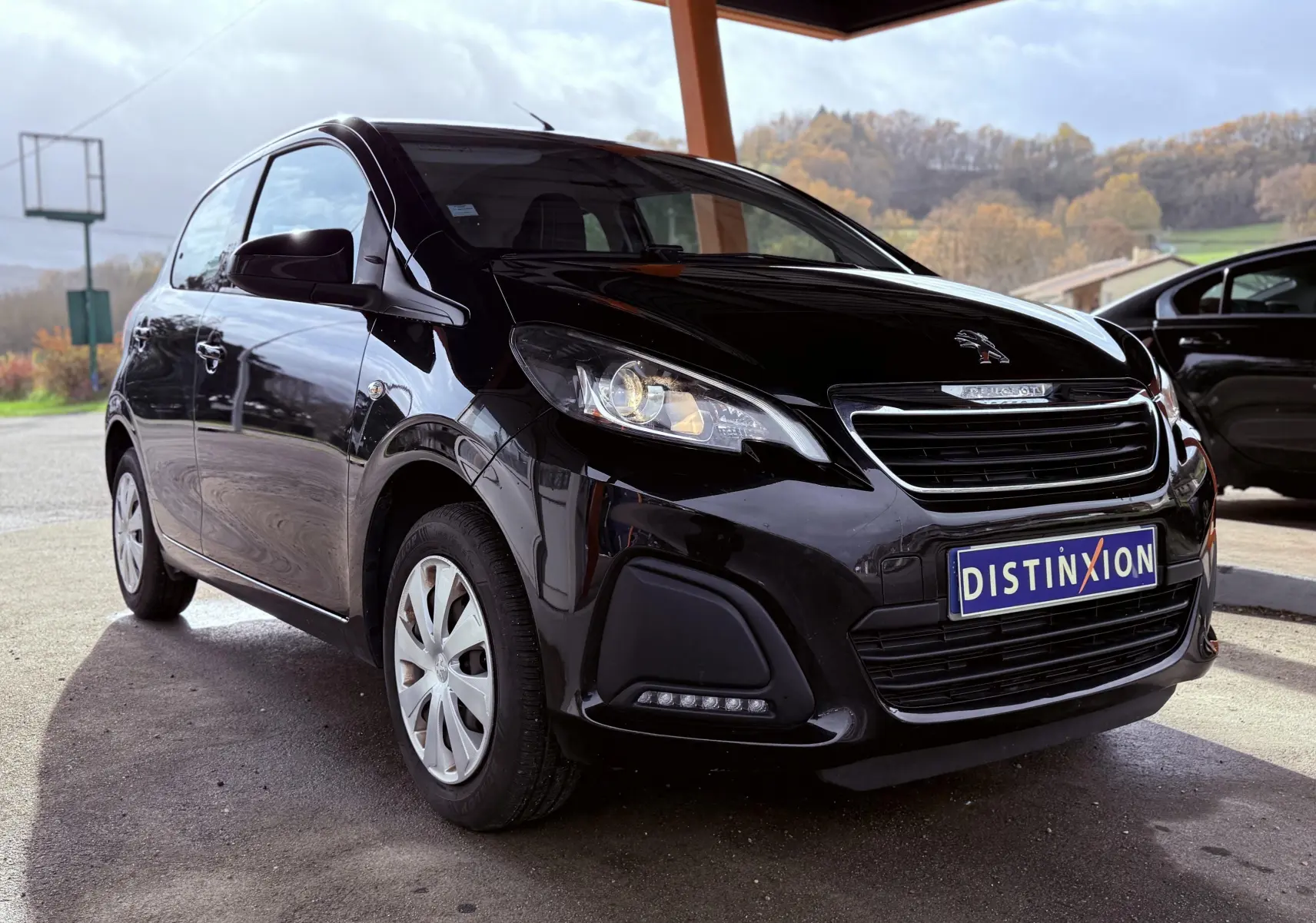 Peugeot 108 noire vue en 3/4 avant droit sous abri avec plaque Distinxion visible sur le pare-chocs.