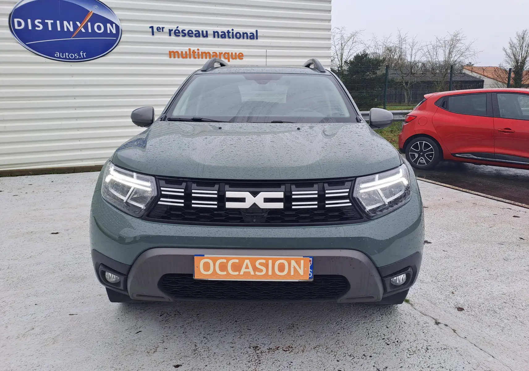 Vue frontale d'un Dacia Duster 2023 kaki Liken avec phares allumés et plaque "OCCASION" visible.