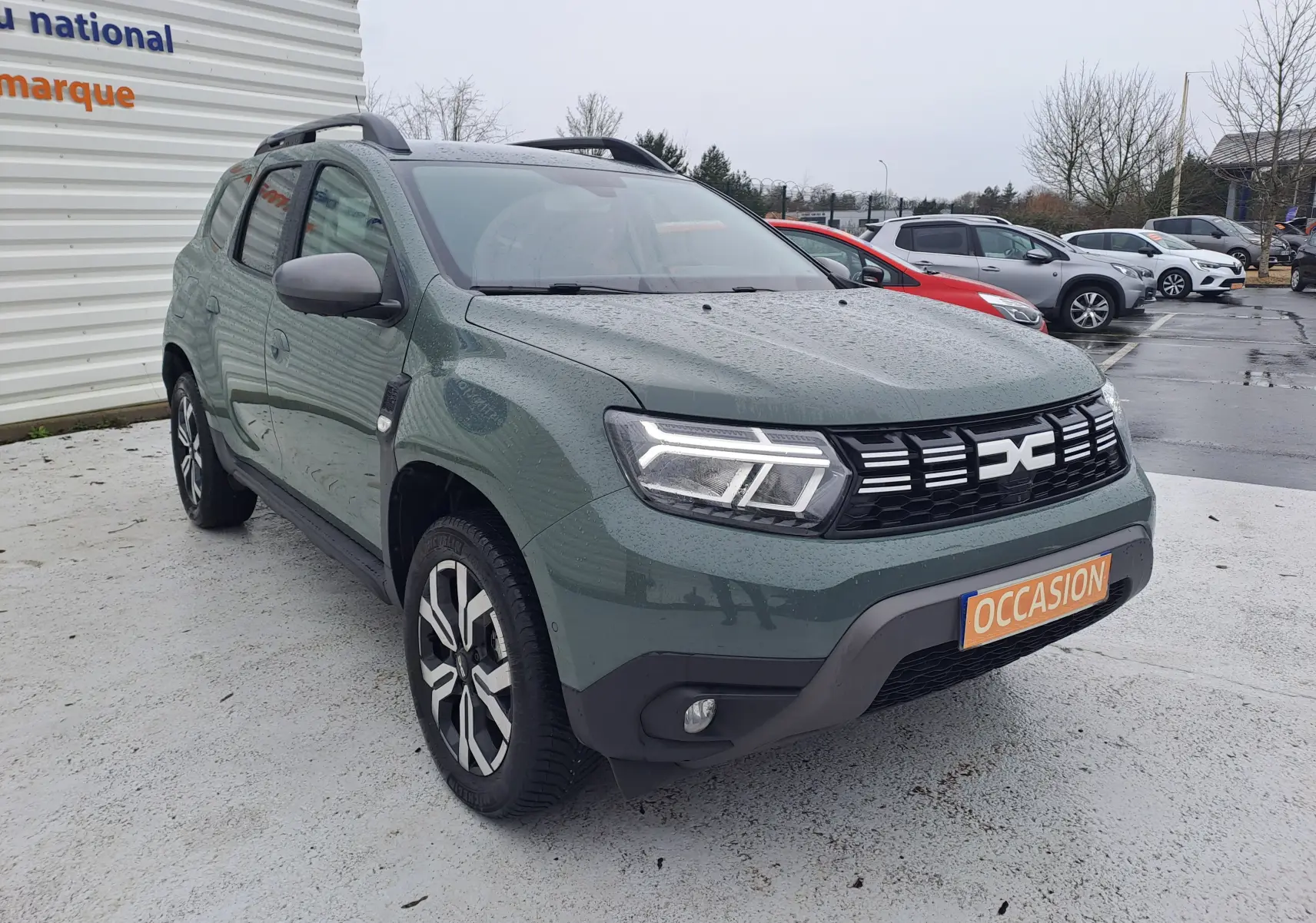 Vue 3/4 avant droit du Dacia Duster 2023 kaki Liken avec calandre noire et jantes bi-ton, stationné sous la pluie.