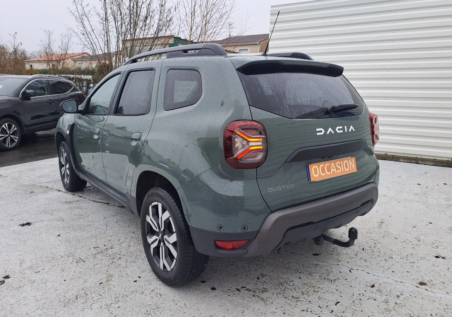 Vue 3/4 arrière droite d'un Dacia Duster 2023 kaki avec attelage et feux arrière allumés sur parking.