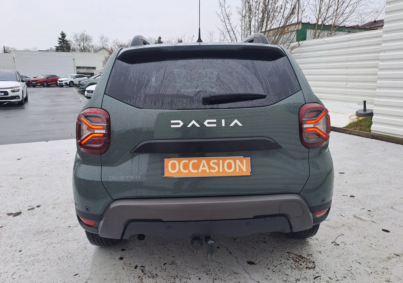 Vue arrière d'un Dacia Duster 2023 kaki Liken avec attelage et feux arrière allumés sur parking humide.