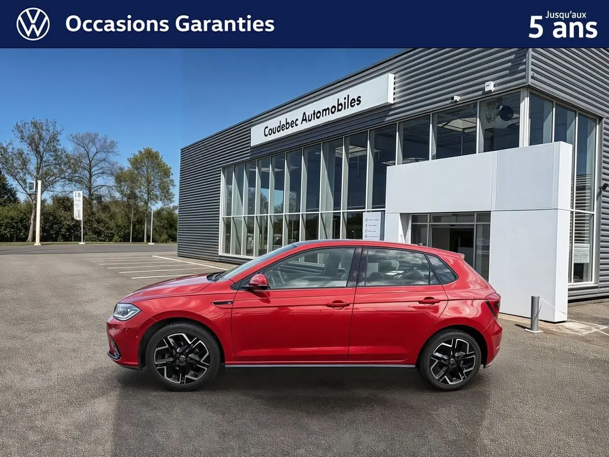 Vue de profil côté gauche d'une Volkswagen Polo rouge métallisée garée devant un bâtiment moderne de concession.