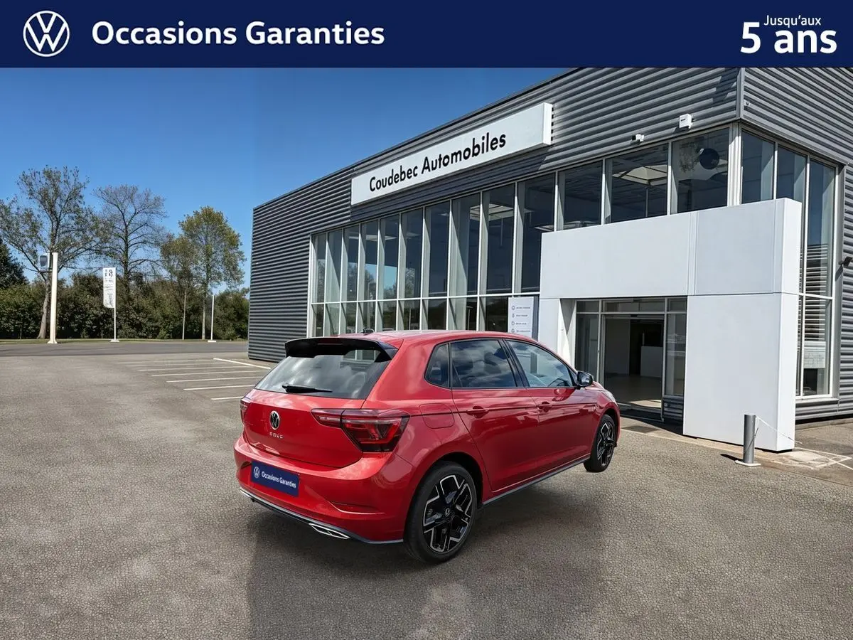 Vue 3/4 arrière droite d'une Volkswagen Polo rouge métallisée garée devant un bâtiment moderne de concession.