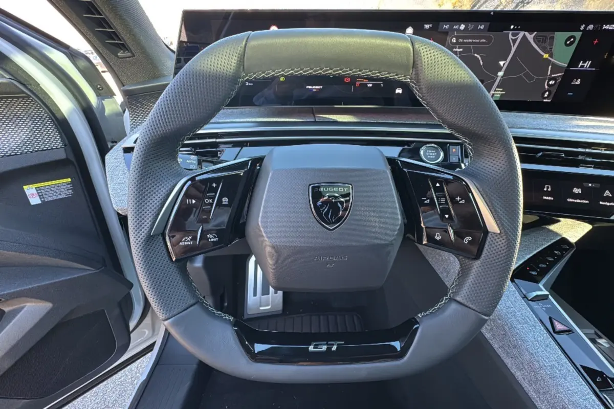 Vue centrée du volant GT noir perforé du Peugeot 3008 Hybrid 2024 avec tableau de bord numérique et portière blanche ouverte.