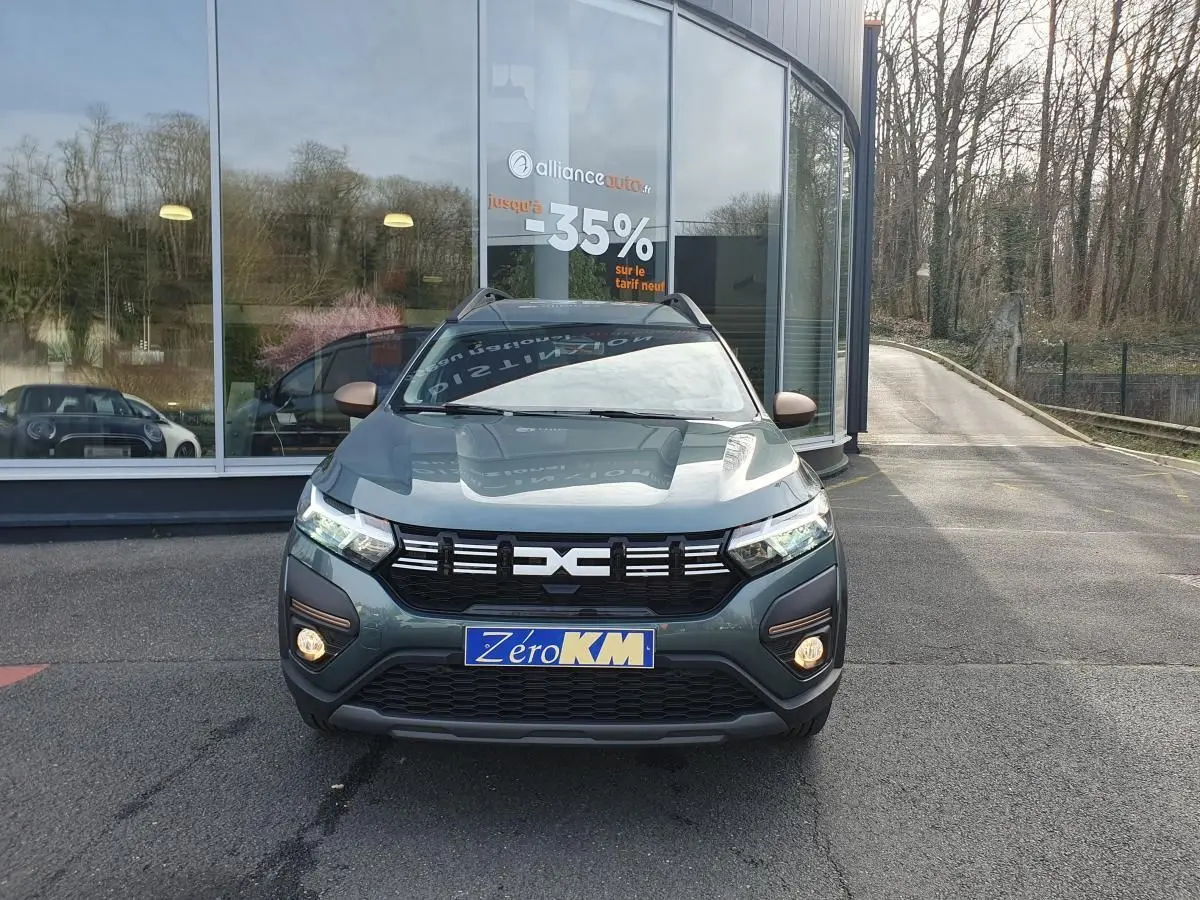 Vue de face du Dacia Jogger vert cèdre métallisé avec calandre noire et feux allumés devant un bâtiment vitré.