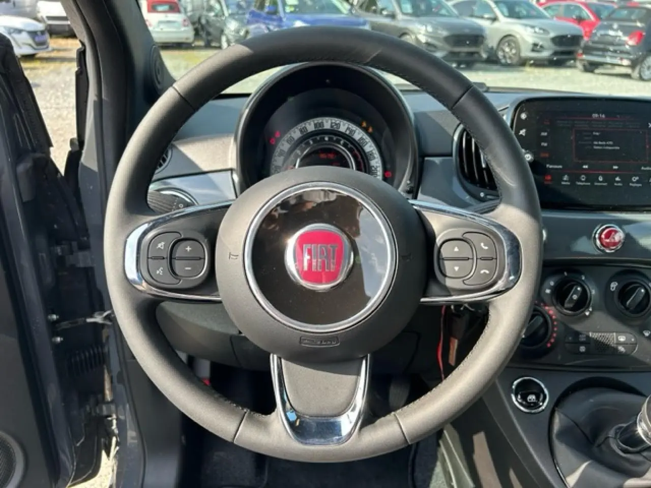 Vue intérieure centrée sur le volant noir avec logo FIAT rouge, tableau de bord et console centrale d'une 500C gris/rouge.