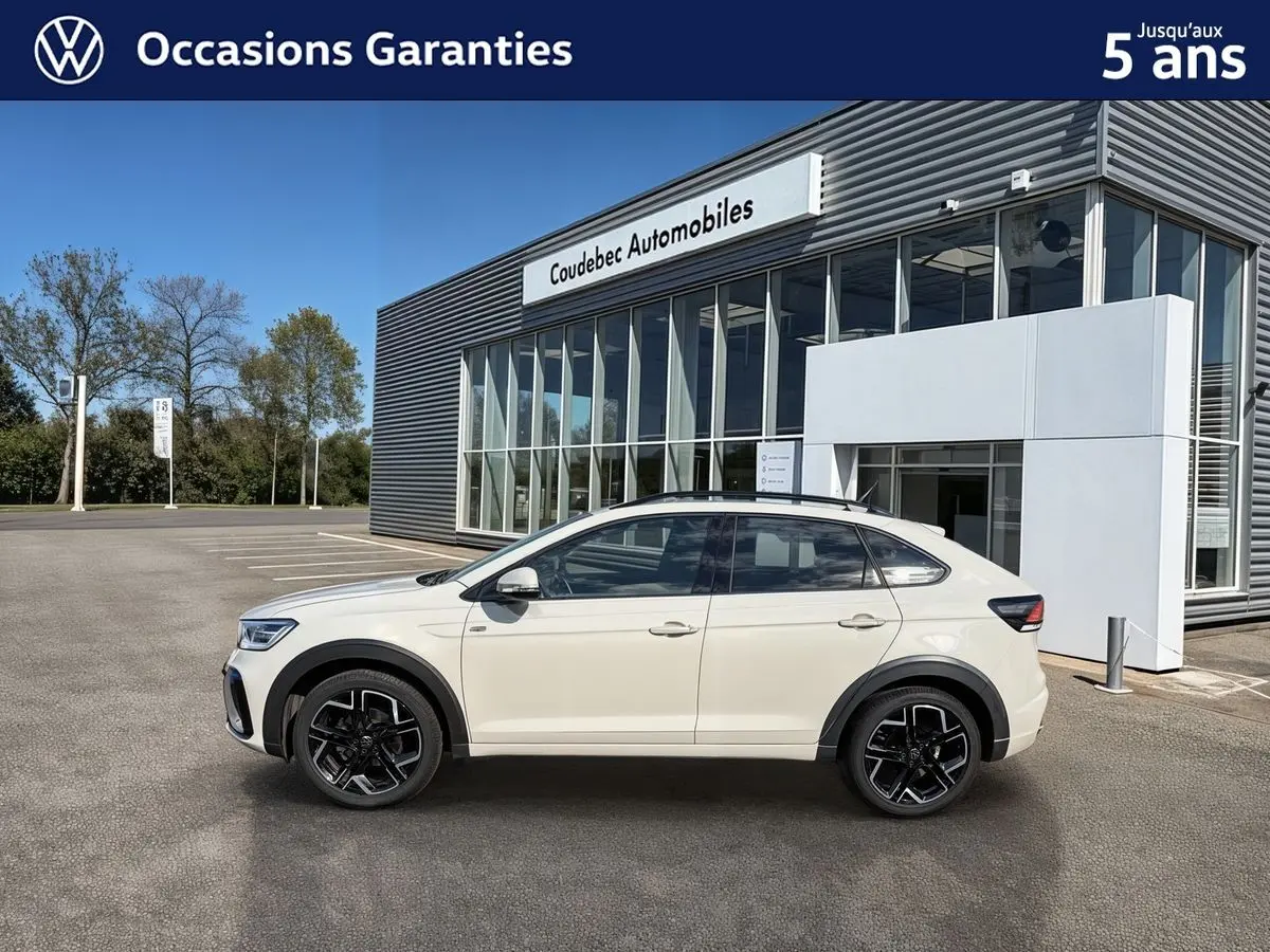 Profil côté gauche du Volkswagen Taigo gris clair garé devant un bâtiment moderne avec grandes baies vitrées.