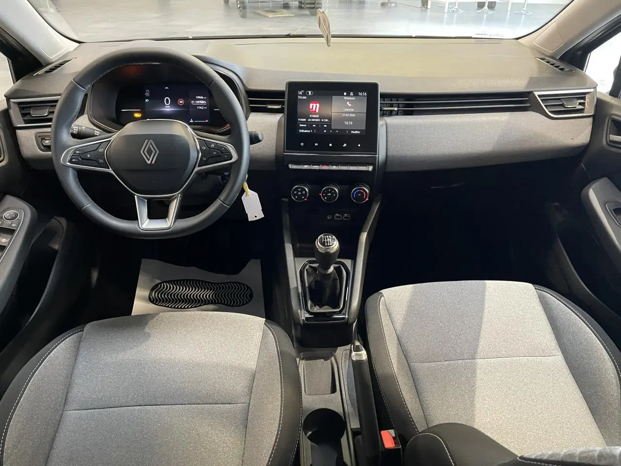Vue intérieure avant de la Renault Clio 2024, tableau de bord noir avec écran tactile et sièges tissu gris.