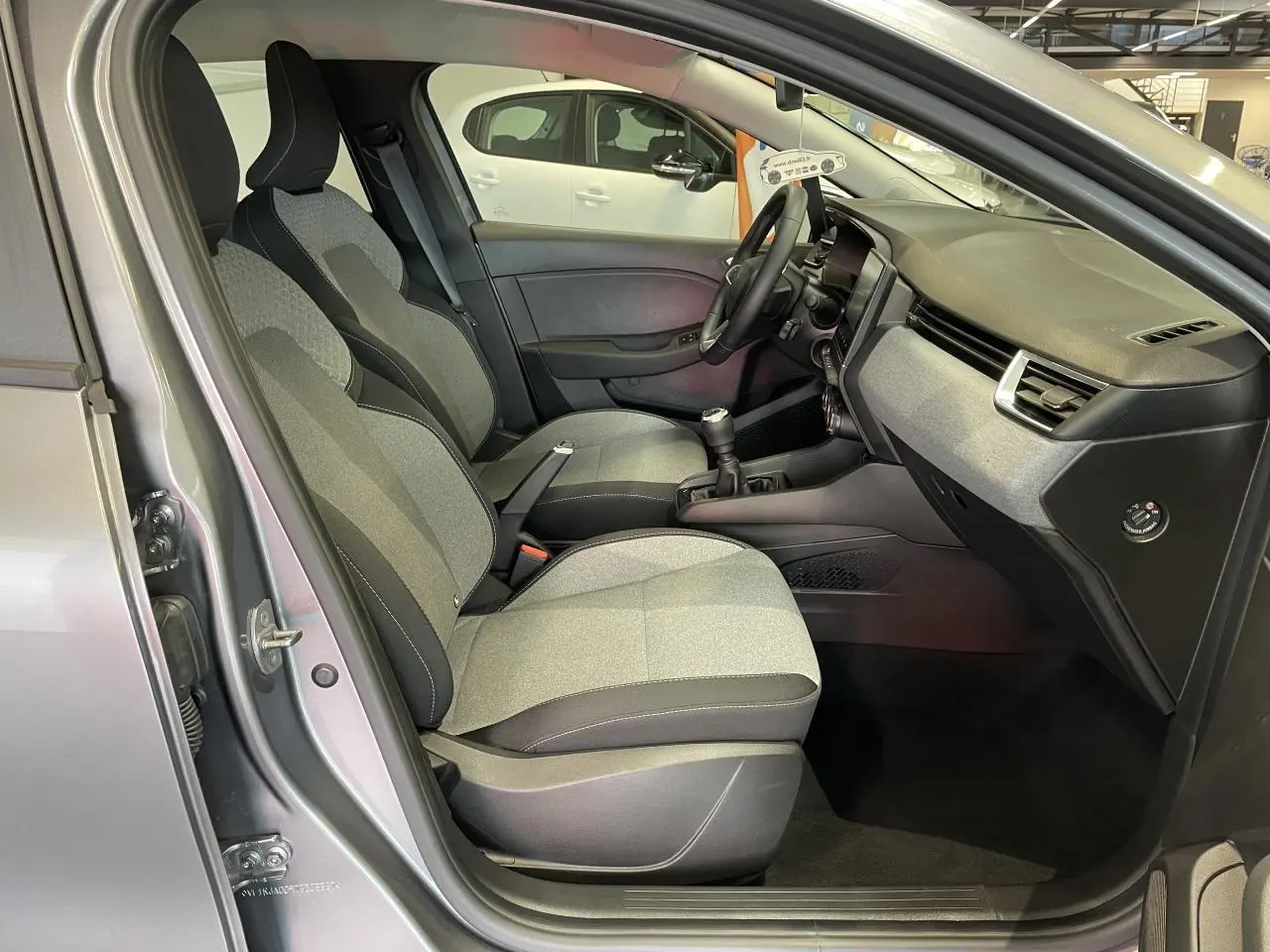 Vue intérieure côté conducteur de la Renault Clio 2024 gris foncé, montrant les sièges tissu gris et la console centrale avec boîte manuelle.