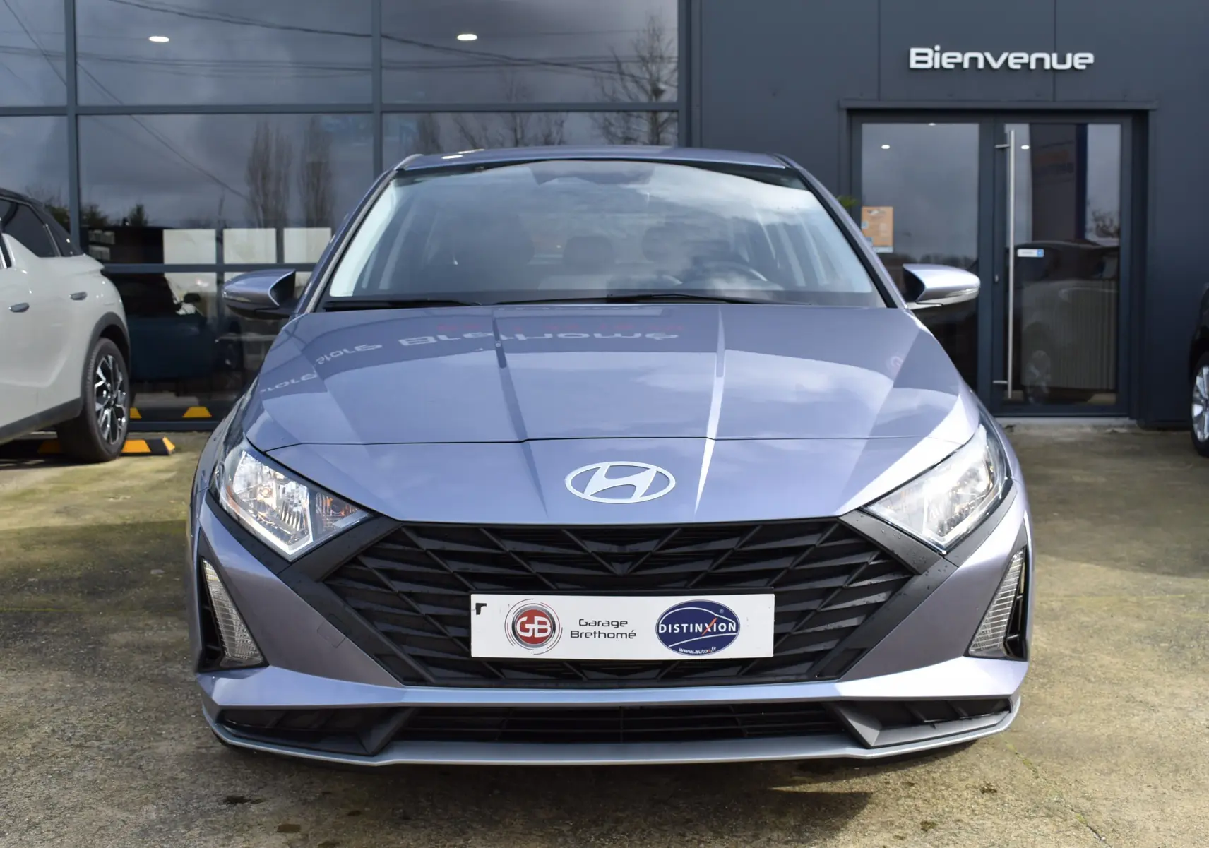 Vue frontale d'une Hyundai i20 bleu clair 2025 avec calandre noire et phares anguleux, devant un bâtiment gris.