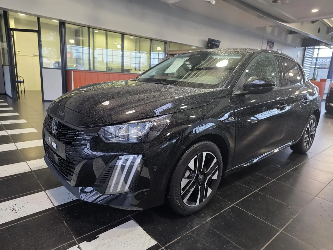 Peugeot 208 hybride noire métallisée vue 3/4 avant droit en intérieur avec jantes alliage bi-tons et feux LED griffés.