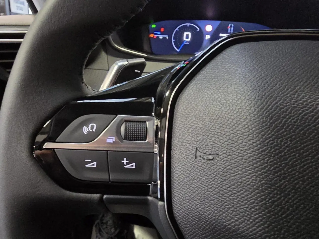 Gros plan sur les commandes gauche du volant cuir noir de la Peugeot 208 hybride 2025 avec tableau de bord numérique.