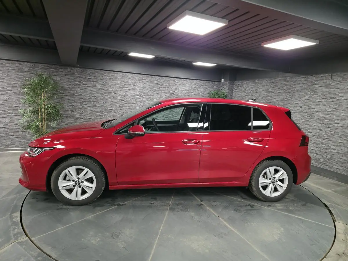 Profil droit d'une Volkswagen Golf VIII rouge métallisée, avec vitres arrière surteintées dans un showroom.