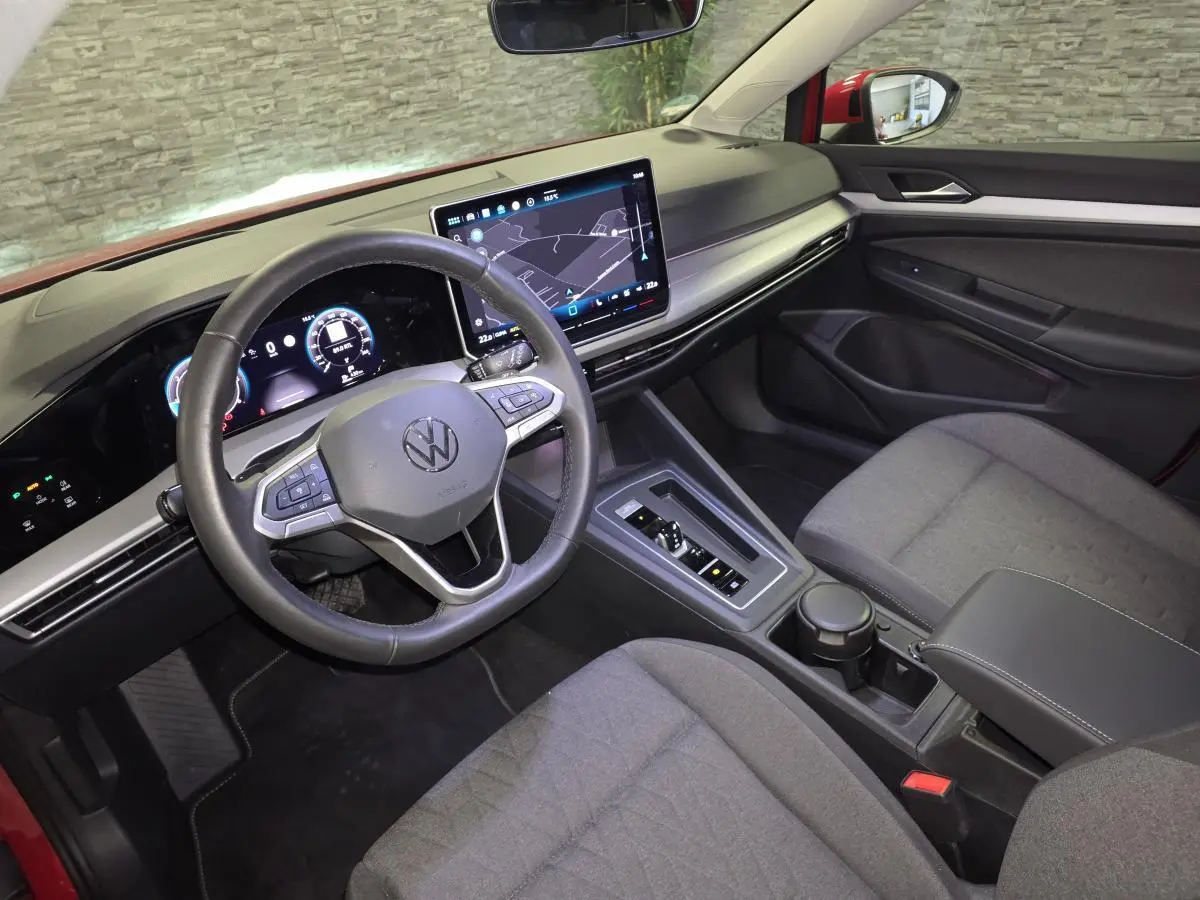 Vue intérieure côté conducteur de la Volkswagen Golf VIII rouge, avec tableau de bord digital et écran tactile central.