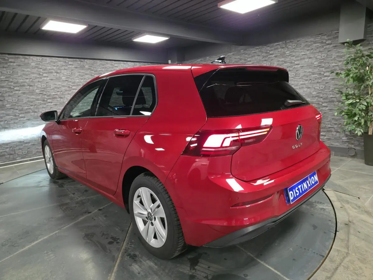 Vue 3/4 arrière droite d'une Volkswagen Golf VIII rouge métallisée dans un showroom avec vitres arrière surteintées.