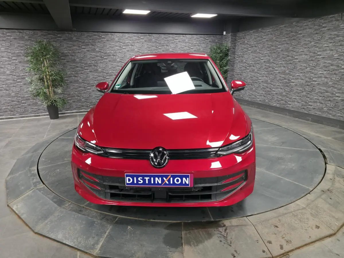 Volkswagen Golf VIII rouge métallisé vue de face dans un showroom avec éclairage intérieur et mur en pierre grise.