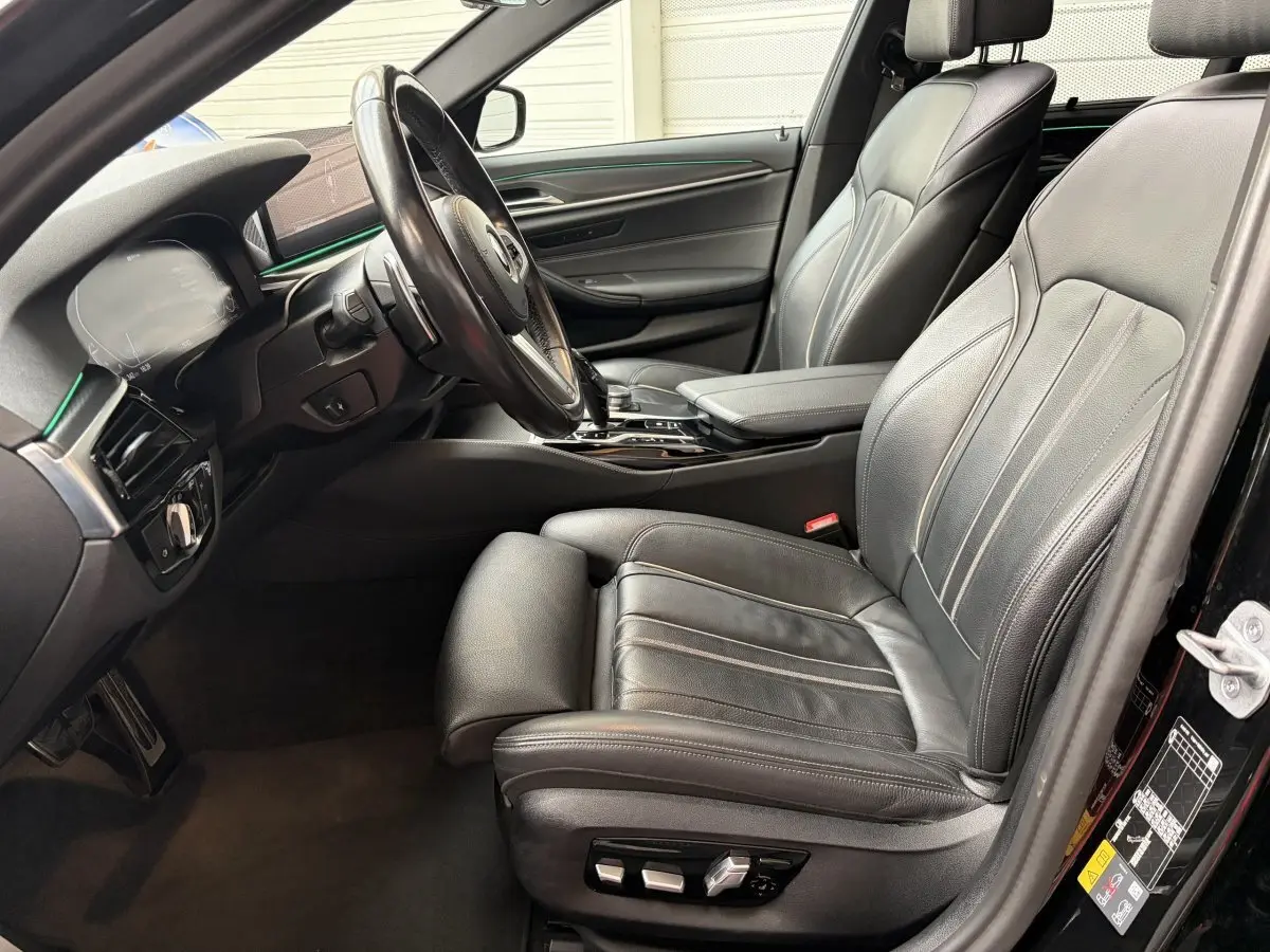 Vue intérieure côté conducteur de la BMW Série 5 Touring 530e noire avec sièges cuir et éclairage d'ambiance vert.