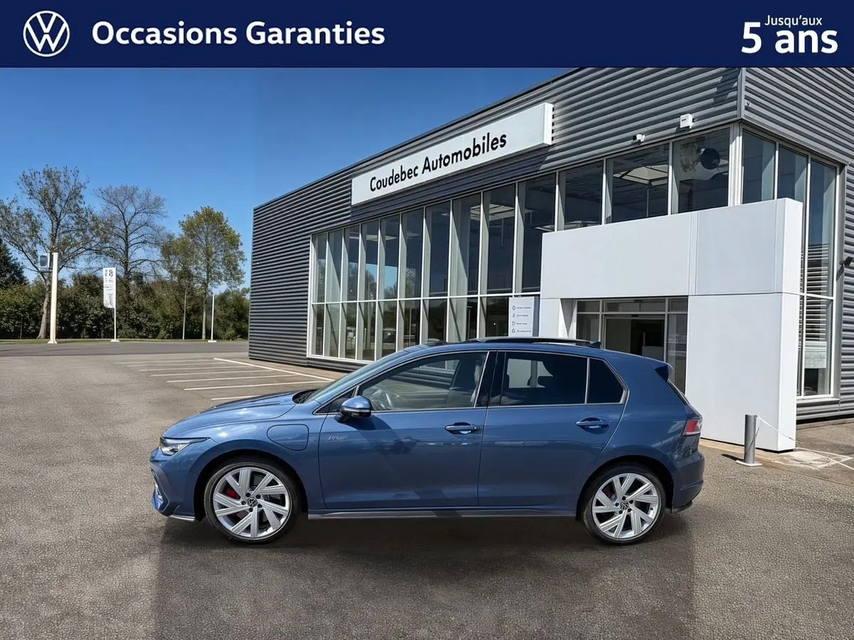 Vue de profil côté gauche d'une Volkswagen Golf 1.5 eHybrid 272 DSG6 bleu métallisé garée devant un bâtiment moderne.