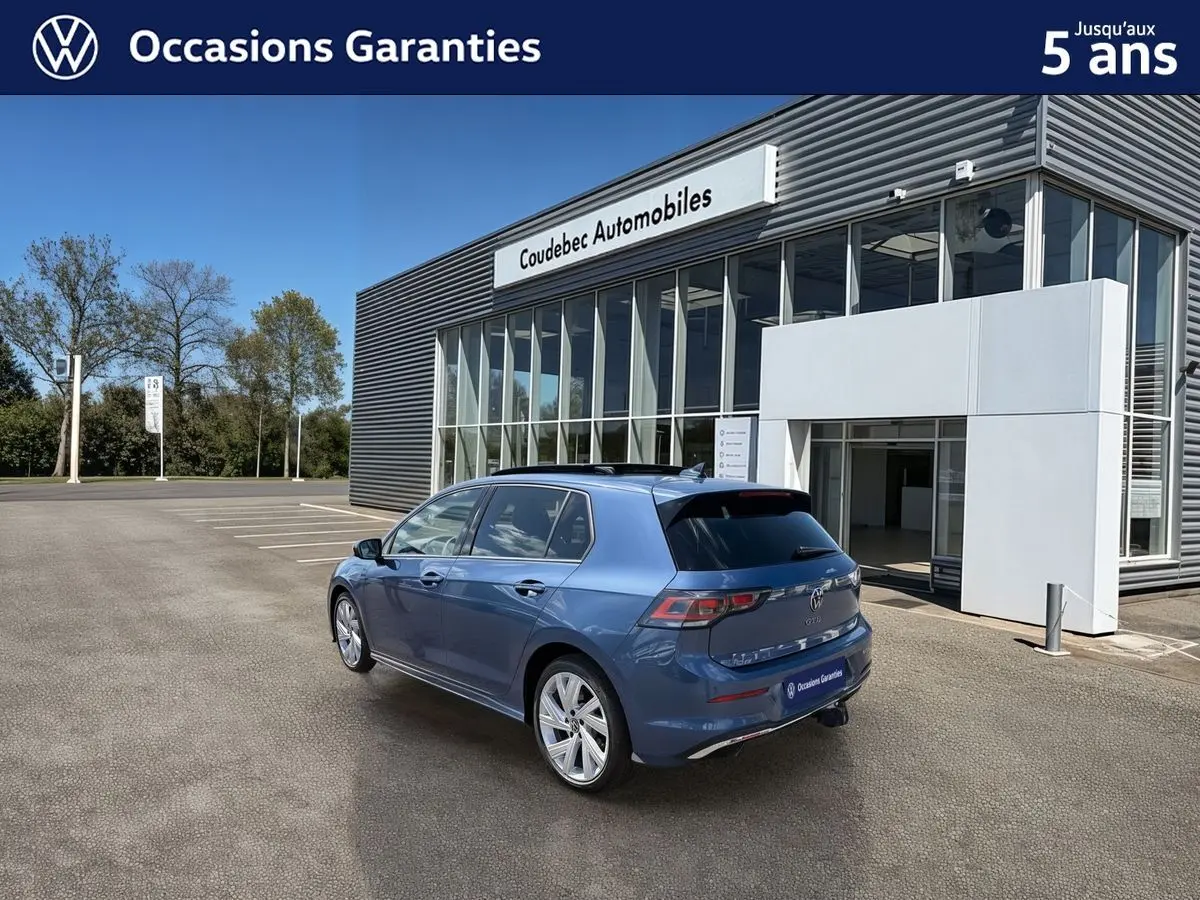 Vue 3/4 arrière droite d'une Volkswagen Golf 1.5 eHybrid 272 DSG6 bleue métallisée, avec toit panoramique et attelage rabattable.