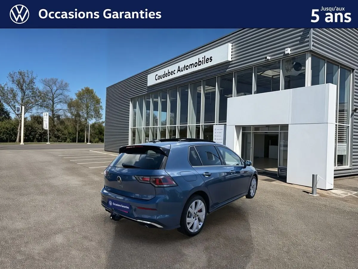 Vue 3/4 arrière droite d'une Volkswagen Golf 1.5 eHybrid 272 DSG6 bleu métallisé avec toit panoramique et attelage rabattable.