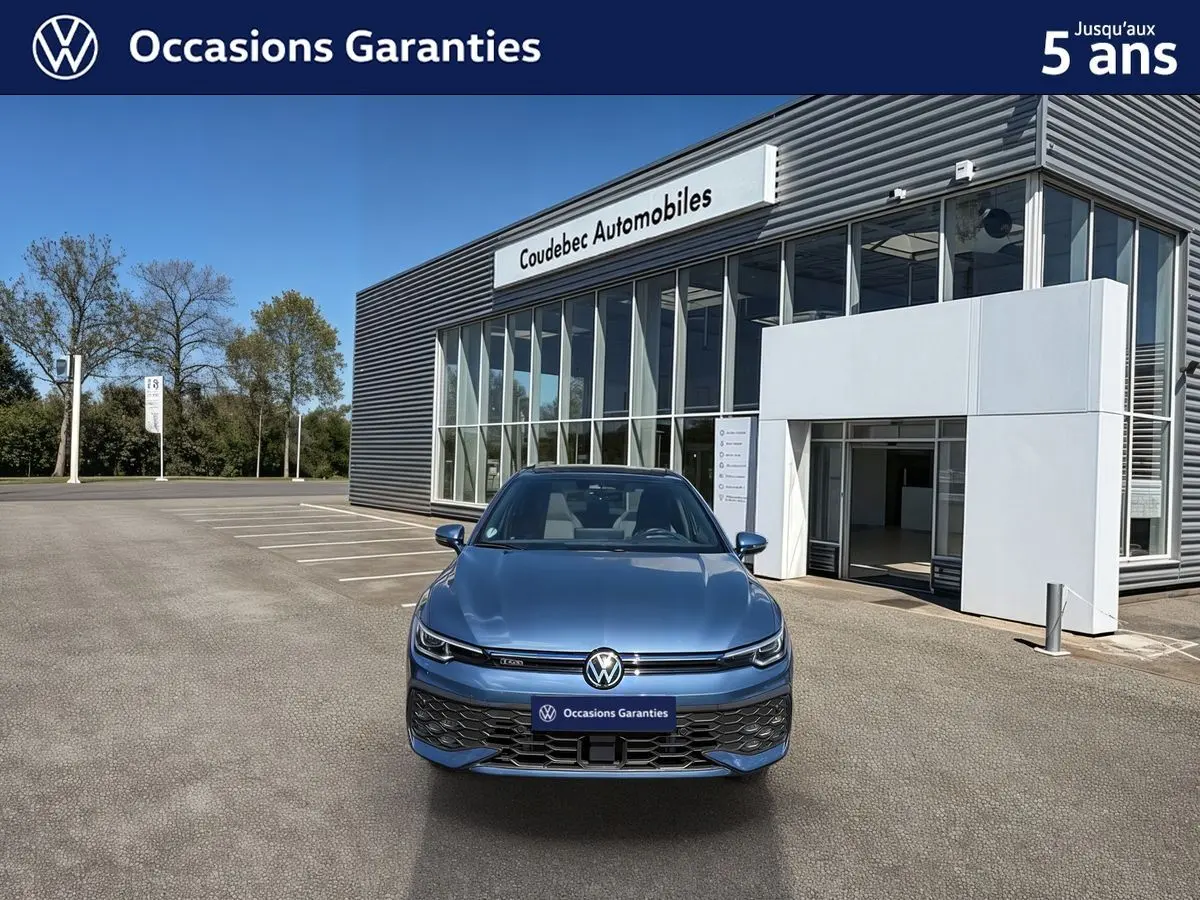 Vue avant d'une Volkswagen Golf 1.5 eHybrid 272 DSG6 bleue métallisée, avec calandre et logo VW visibles, devant un showroom.