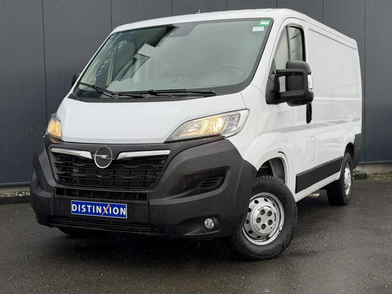 Vue 3/4 avant droit d’un Opel Movano Fourgon blanc avec pare-chocs noir et phares allumés sous la pluie.