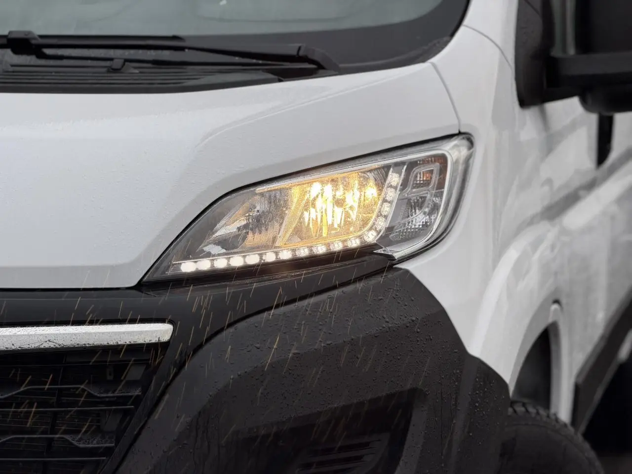 Gros plan sur le phare avant droit allumé d’un Opel Movano Fourgon blanc sous la pluie.