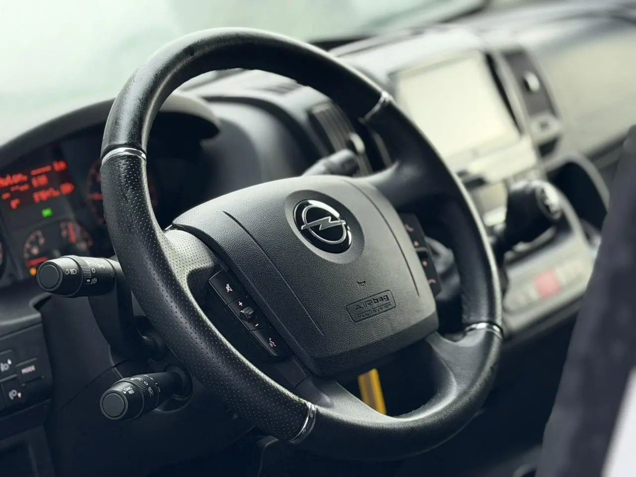 Gros plan sur le volant en cuir noir de l’Opel Movano Fourgon 2024 avec tableau de bord et écran tactile flou en arrière-plan.