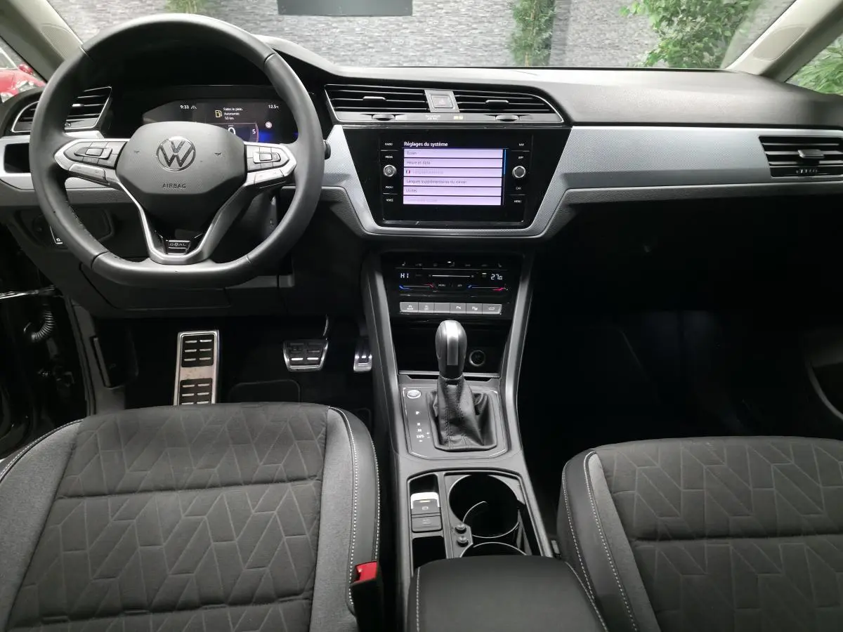Vue intérieure avant du Volkswagen Touran 2025 noir, montrant le volant multifonction, l'écran tactile central et la console avec boîte auto.