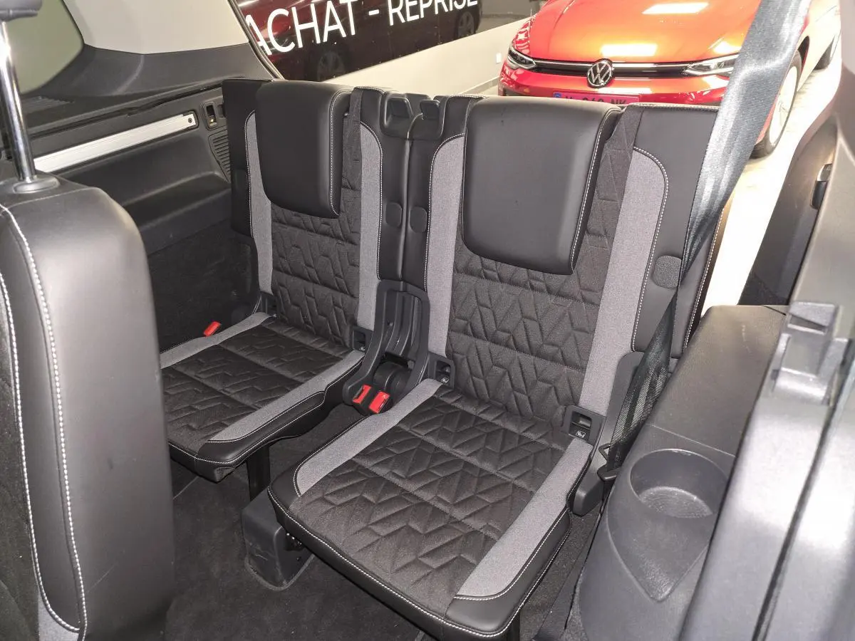 Vue intérieure sur la troisième rangée de sièges noirs et gris du Volkswagen Touran 2025, avec arrière d’un autre véhicule rouge visible.