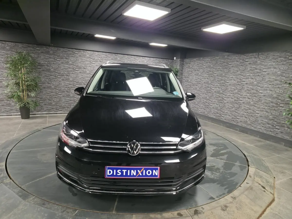 Vue avant d'un Volkswagen Touran noir nacré 2025 avec calandre chromée et plaque Distinxion en intérieur showroom.