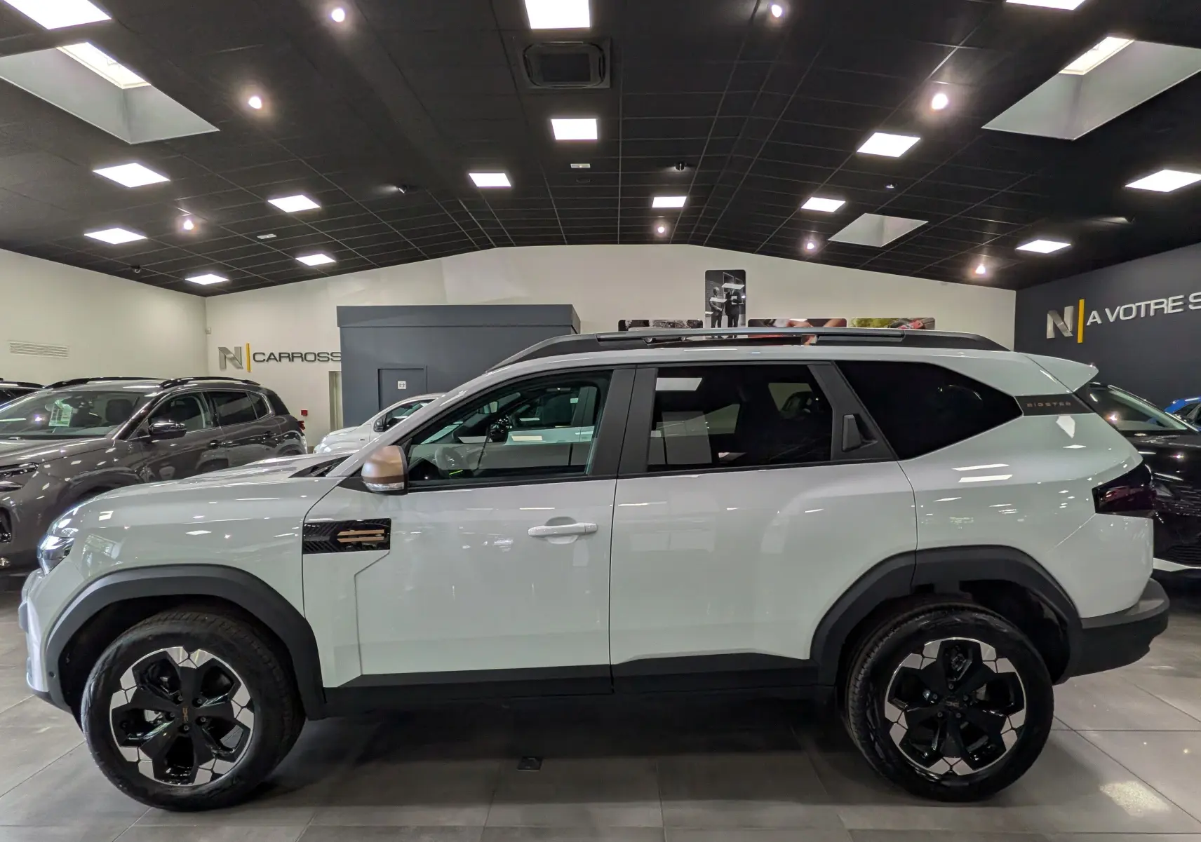 Profil droit du Dacia Bigster blanc Glacier 2026 avec jantes noires et toit ouvrant en showroom.