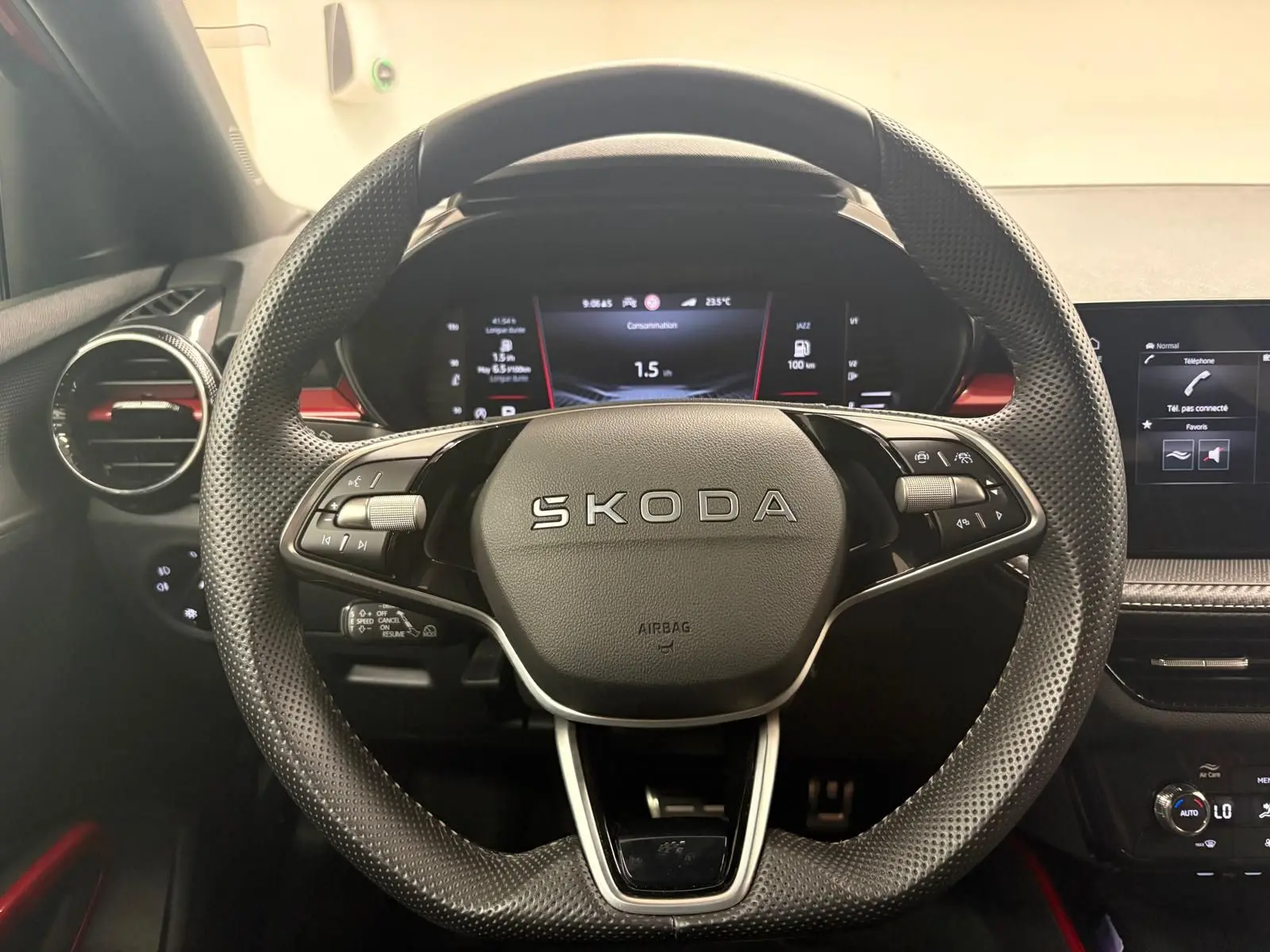 Vue rapprochée du volant perforé noir avec logo SKODA, tableau de bord numérique et détails rouges intérieurs visibles.