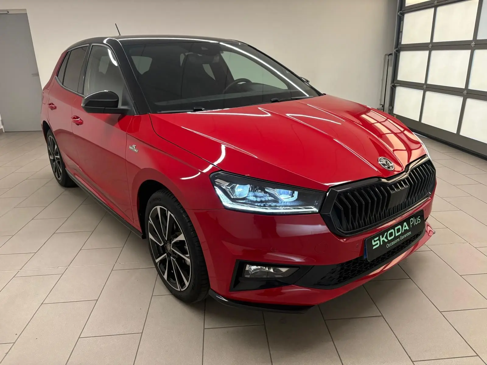 Vue 3/4 avant gauche d'une Skoda Fabia Monte Carlo rouge Velvet avec toit noir et phares allumés en intérieur.