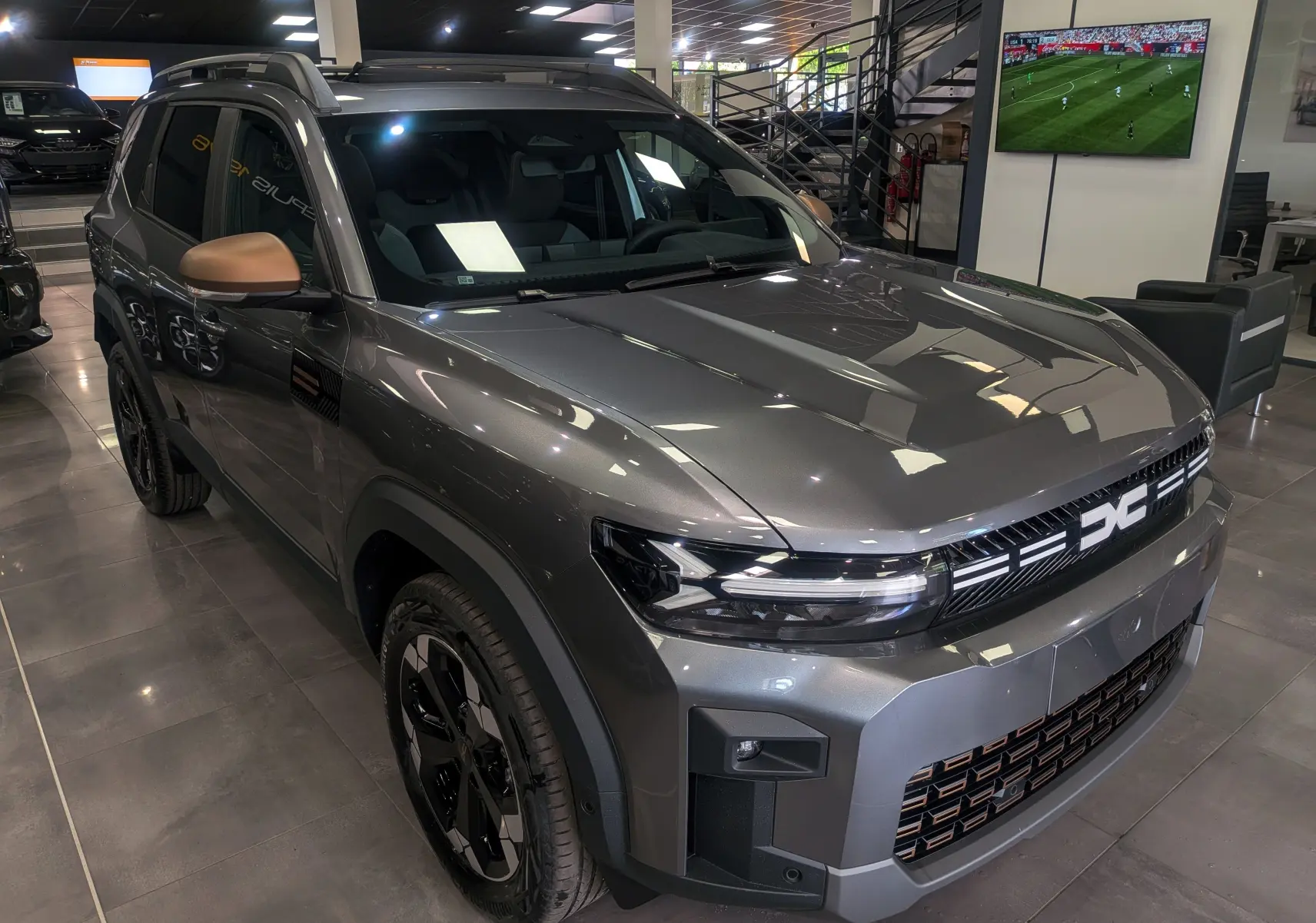 Vue 3/4 avant droit du Dacia Bigster gris schiste avec détails noirs et rétroviseurs couleur cuivre en showroom.