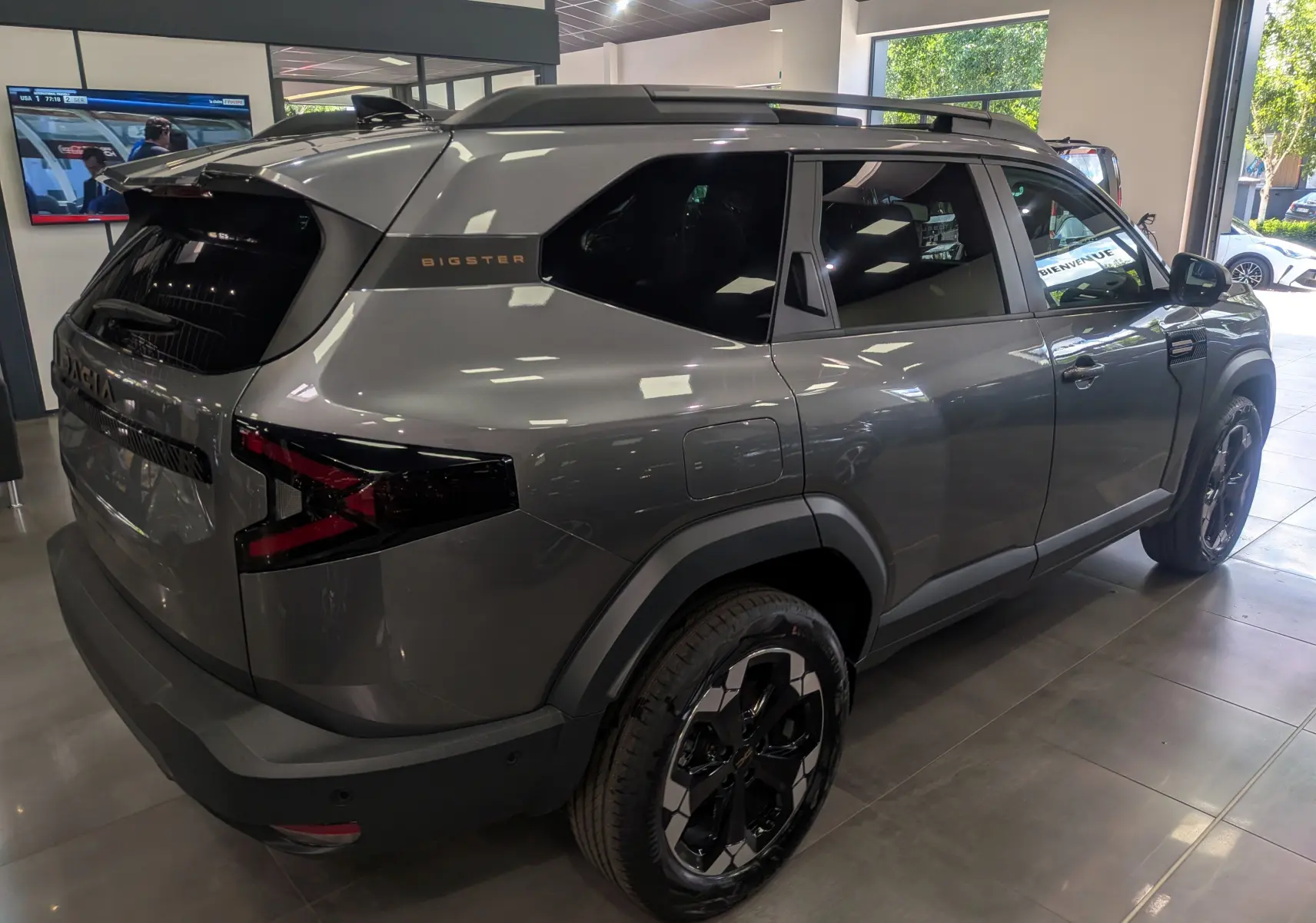 Vue 3/4 arrière droite du Dacia Bigster gris schiste avec jantes noires et toit avec barres de toit en concession.