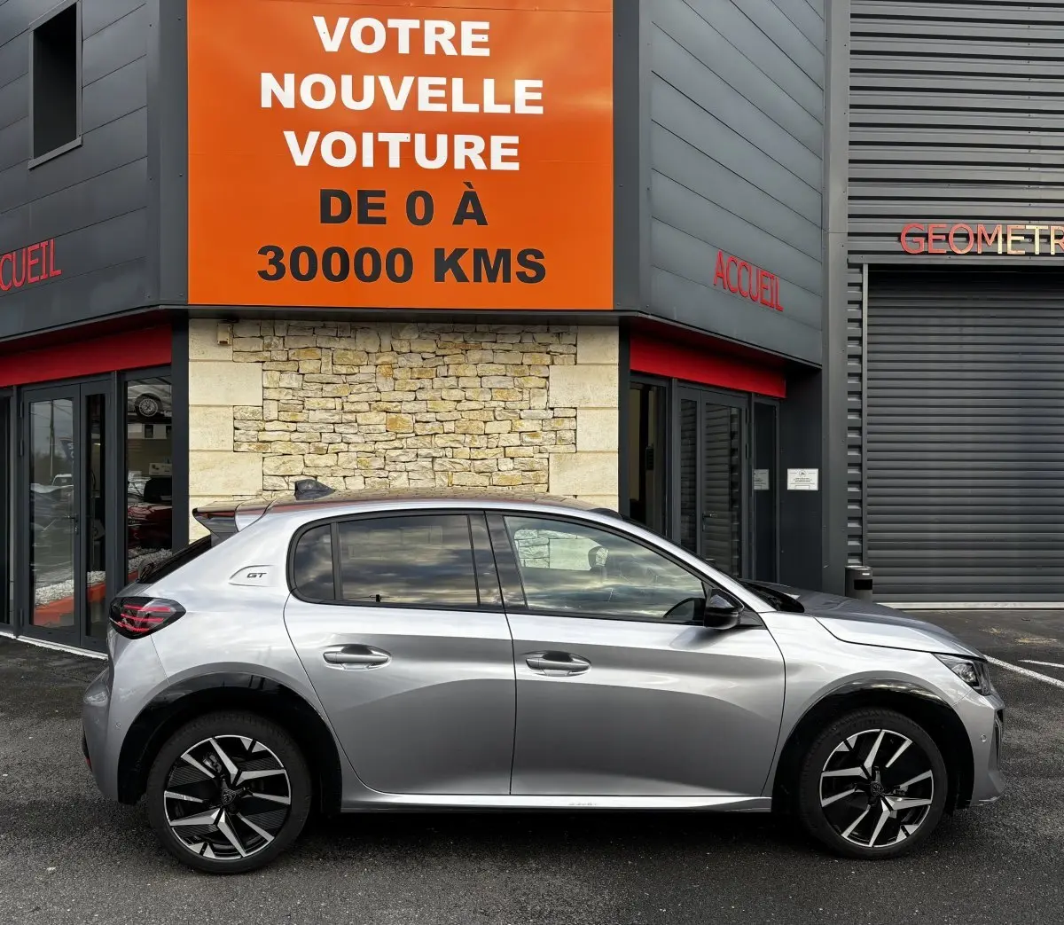 Vue de profil côté gauche d'une Peugeot 208 Hybrid 110 e-DCS6 GT gris clair garée devant un bâtiment moderne.